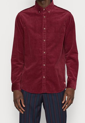 Mężczyzna ubrany w bordową, zapinaną na guziki koszulę ze sztruksu z długimi rękawami oraz granatowe spodnie z czerwonymi pionowymi paskami.