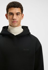 Felpa nera con cappuccio grande, caratterizzata da un logo "BOSS" in rilievo tono su tono. Indossata sopra un dolcevita crema, che mostra una superficie liscia.