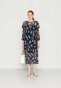 Vestido floral azul-marinho com tecido transparente, mangas com volantes e comprimento midi. Combinado com sapatos de salto finos e uma carteira branca.