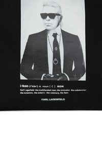 Crni materijal s nijansama sive na kojem se nalazi slika osobe u odijelu s naočalama, uz tekst koji definira "i-kon" i referencira Karla Lagerfelda.