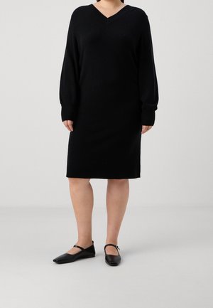 Femme portant une robe en maille noire à manches longues avec un décolleté en V et des ballerines Mary Jane noires, debout sur un fond clair uni.