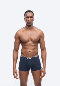 Μυώδης άντρας χωρίς πουκάμισο, όρθιος με το πρόσωπο προς τα εμπρός, φοράει μπλε ναυτικό boxer briefs με το λογότυπο BOSS, σε απλό ανοιχτό φόντο.