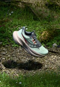Chaussure de sport vert clair avec des accents gris, tige texturée, rayures rouges et noires, et une semelle épaisse et rembourrée, suspendue au-dessus d'un fond naturel.