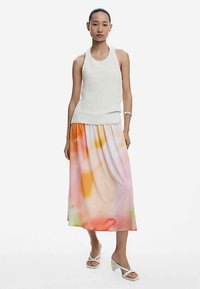 Fehér bordázott ujjatlan felső, légies, pasztellszínű maxi szoknyával, amely absztrakt mintákat visel. A modell fehér pántos szandált visel.