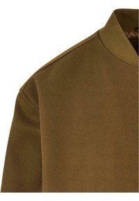 Épaule et col d'une veste en laine marron avec couture visible et col ras du cou côtelé, présenté sur fond blanc.