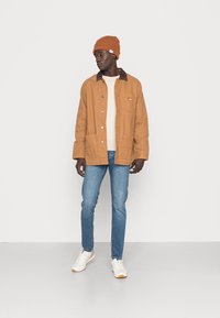 Braune Baumwolljacke mit dunklem Kragen, beiges Hemd, blaue Jeans und weiße Sneakers, abgerundet mit einer orangen Strickmütze.