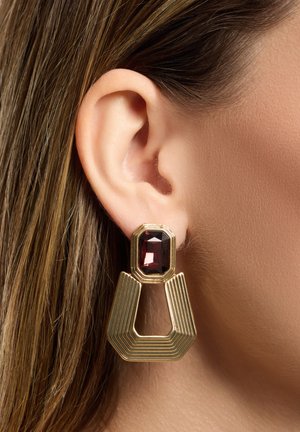 Goldfarbene Ohrringe mit einem rechteckigen roten Edelstein auf einem strukturierten geometrischen Design. Die Ohrringe sind groß und haben eine geschichtete Form.