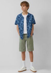 Kurzmanteliges blaues Hemd mit Blattmuster, kombiniert mit olivgrünen Shorts. Weißes T-Shirt, weiße Socken und cremefarbene Sneaker.