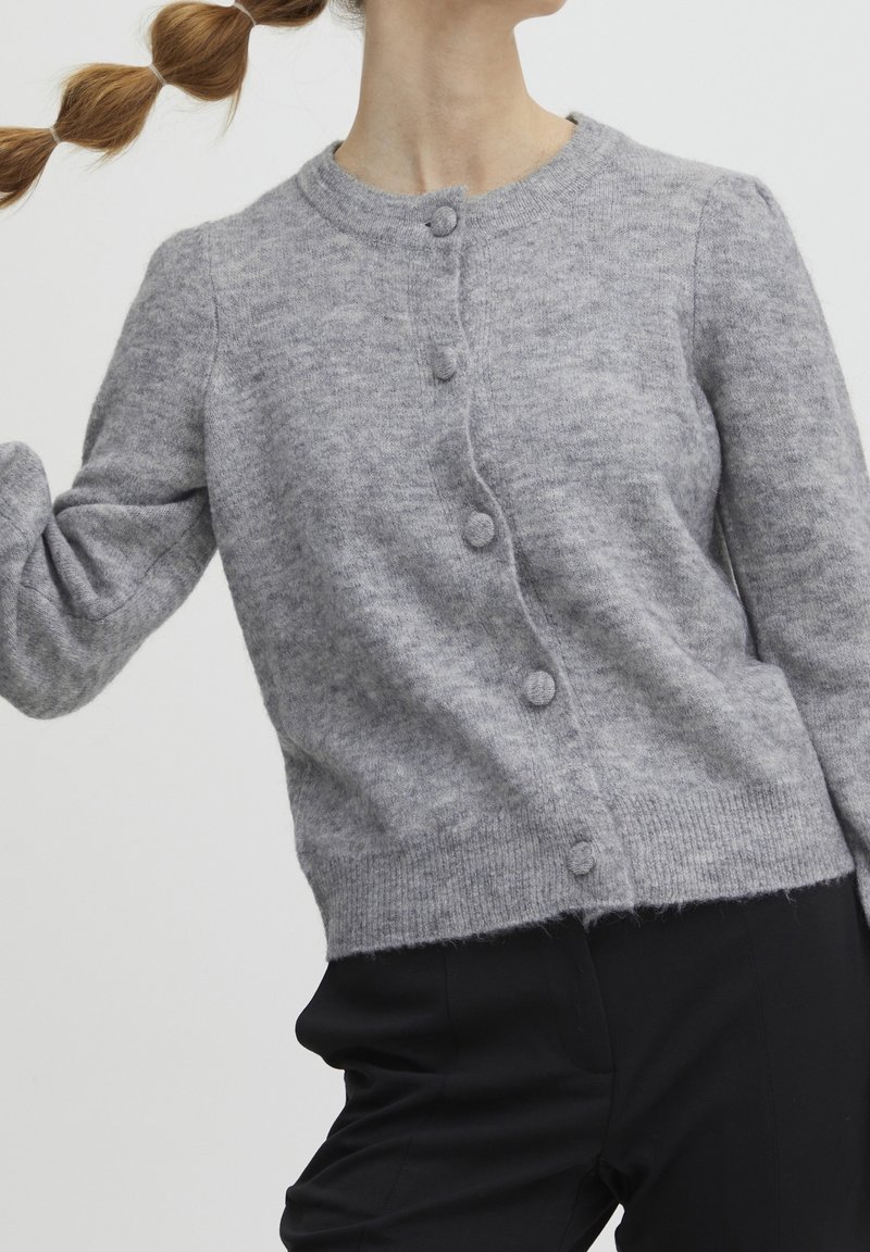 Femme portant un cardigan gris boutonné en tissu texturé et un pantalon noir, les cheveux coiffés en tresse bulle sur un fond uni.
