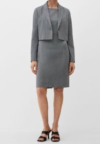 Harmaa räätälöity bleiseri, jossa on lyhyt leikkaus, harmaa istuva mekko ja mustat korokepohjaiset sandaalit. Sujuva tekstiili, hienovarainen kuvio ja yksittäinen nappi yksityiskohtana.