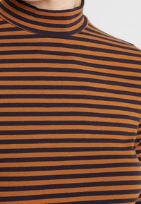Acercamiento de una camiseta de cuello alto con rayas horizontales en mostaza y azul marino oscuro en la parte superior del torso y el cuello de una persona.