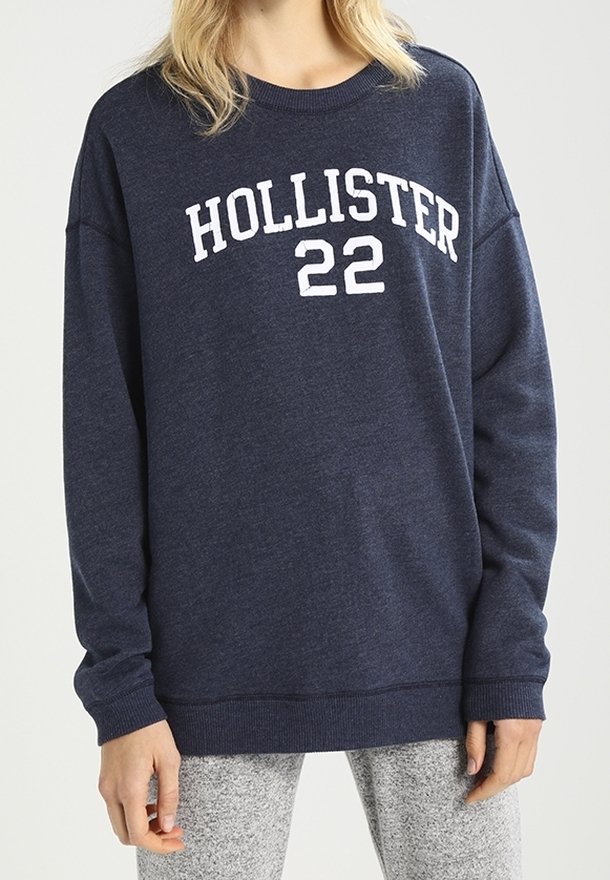 Sweat-shirt bleu marine Hollister avec texte blanc "HOLLISTER 22" porté par une personne, associé à un pantalon gris clair.