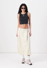 Black cropped tank top with white logo; cream-colored, pleated maxi skirt; gray and white sneakers with visible laces and socks.