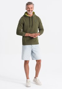 Sudadera color verde oliva con un forro interior amarillo, cordones y detalles laterales, combinada con pantalones cortos de mezclilla claros y zapatillas blancas.