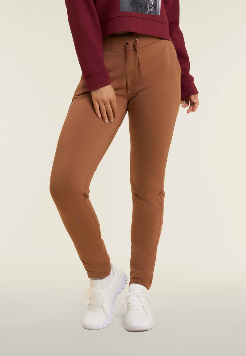 Pantaloni della tuta marroni a tapered con vita elasticizzata e dettagli con coulisse, abbinati a sneakers bianche. Tessuto liscio e aderente.