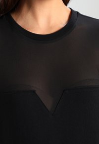 Top nero con sovrapposizione trasparente, scollo rotondo e motivo a V sul petto. Il tessuto sembra leggero e strutturato.