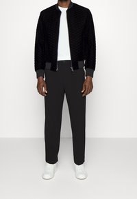 Svart vadderad bomberjacka med grå detaljer, kombinerad med svarta byxor och vita sneakers. Visad mot en vit bakgrund.