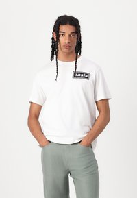 White cotton T-shirt with a black rectangular "basis" graphic on the left chest, worn with light green pants. Relaxed fit, short sleeves.