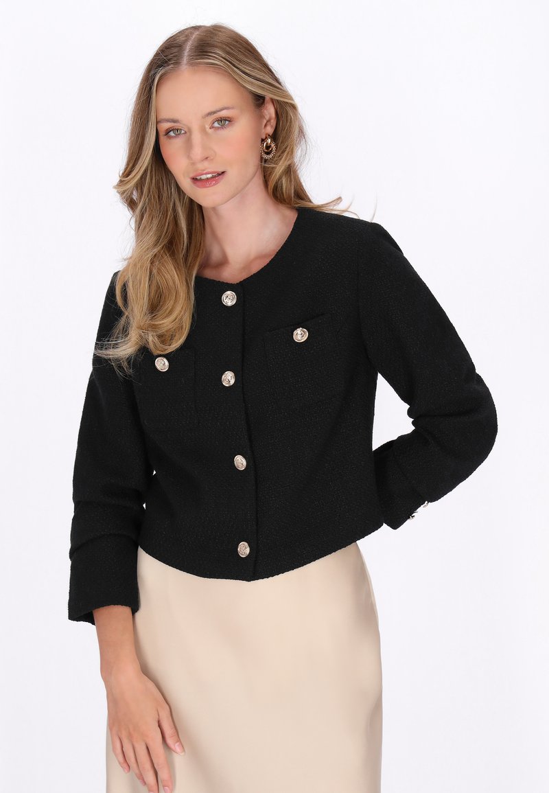 Blonde Frau trägt eine schwarze Jacke mit goldenen Knöpfen und einen beigen Rock, steht vor einem schlichten weißen Hintergrund.