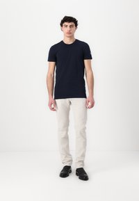 Námořnické modré tričko s krátkým rukávem z bavlněného materiálu, spárované se světle béžovými kalhotami a černými slip-on botami, vystavené na neutrálním pozadí.