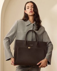 Brown textured leather handbag with dual handles, gold zipper accents, and a front pocket. Styled with a gray tailored outfit.