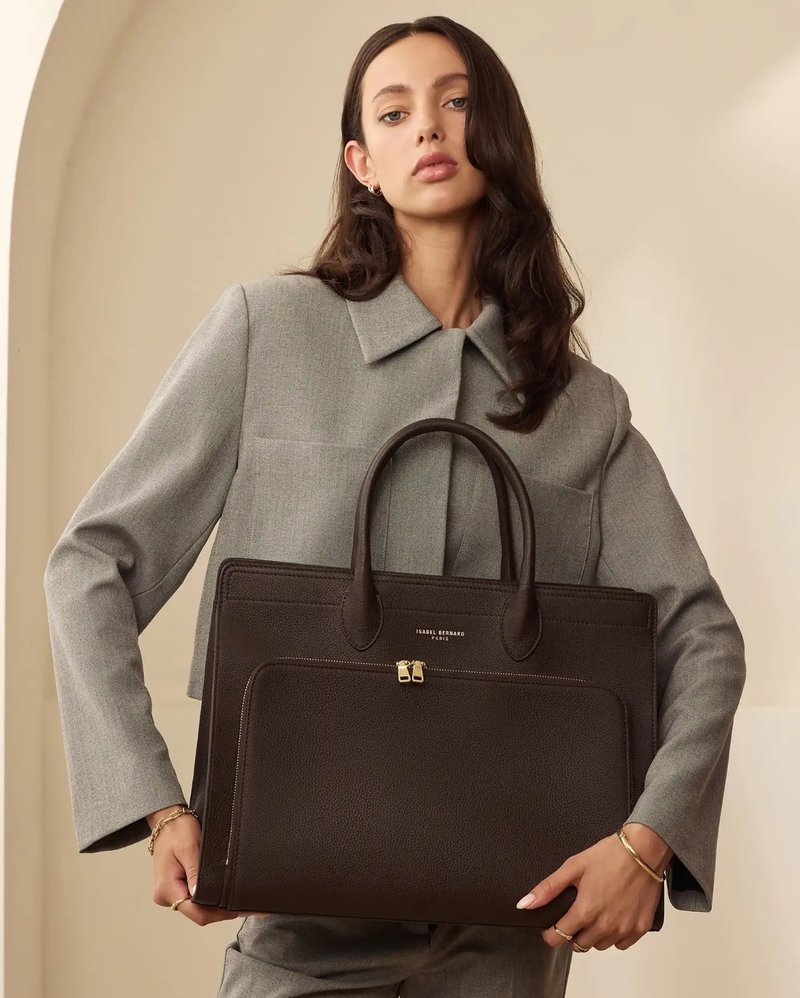 Brown textured leather handbag with dual handles, gold zipper accents, and a front pocket. Styled with a gray tailored outfit.