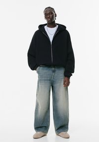 Crna zip-up dukserica, bijela majica ispod, svijetloplave široke traperice s izblijedjelim dijelovima i bež tenisice. Model stoji naslonjen na bijelu pozadinu.