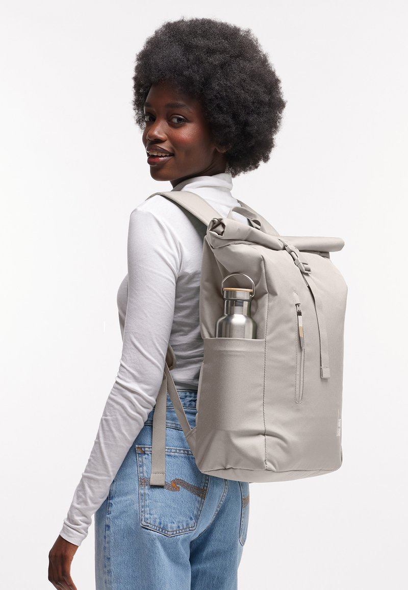 Woman wearing a light gray backpack holding a stainless steel water bottle in side pocket, facing sideways with a slight smile.