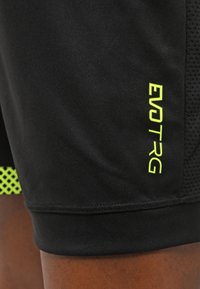 Shorts de sport noirs avec le logo "EVOTRG" jaune néon sur le côté inférieur, portés sur la cuisse d'une personne à la peau brune visible.