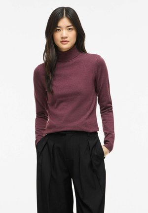 Jeune femme aux longs cheveux foncés portant un pull à col roulé bordeaux et un pantalon plissé noir, les mains dans les poches.