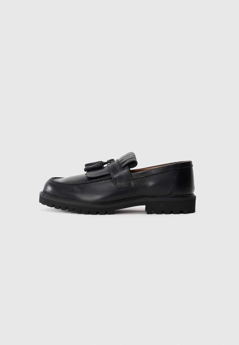 Mocassin en cuir noir avec des glands, bout arrondi et semelle épaisse à crampons, présenté en profil sur un fond clair uni.