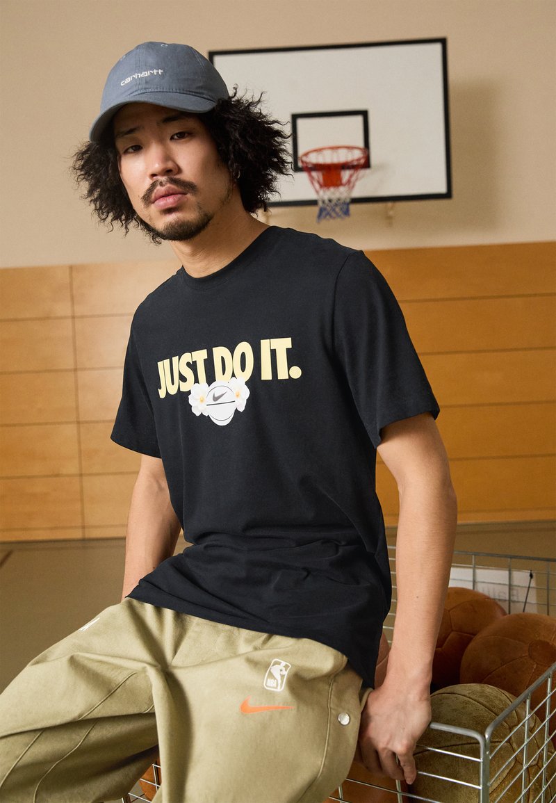 Svart bomulls T-skjorte med teksten "JUST DO IT." og blomsterdesign, kombinert med olivengrønne oversized bukser med NBA-logo og oransje Nike-detalj.