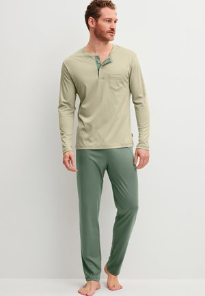 Uomo in piedi a piedi nudi che indossa una maglia henley beige a maniche lunghe e pantaloni verdi dal taglio morbido su uno sfondo chiaro e uniforme.