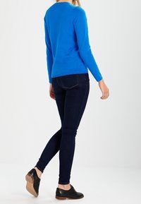 Niebieski sweter z długim rękawem o dopasowanym kroju, w połączeniu z ciemnoniebieskimi spodniami typu slim fit. Buty są czarne, niskoprofilowe i o minimalnych detalach.