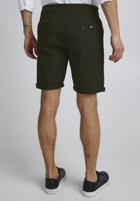 Shorts vert foncé avec des ourlets retroussés, fabriqués à partir d'un tissu lisse. Équipés de poches arrière avec des fermetures à bouton et d'un détail rayé discret.