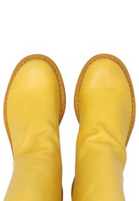 Gele leren kniehoge laarzen met een gladde textuur, ronde neus en contrasterende tan stiksels rondom de zool. Zachte, slordige uitstraling.
