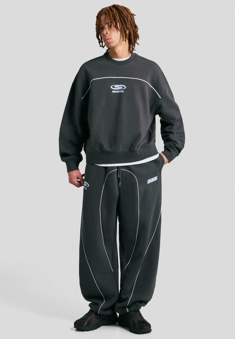 Temno siva trenerka z ujemajočimi se hlačami, ki imata bele obrobe in logotipe, narejena iz mehkega materiala z udobnim krojem in širokimi nogavicami.