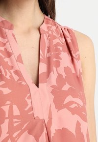 Ärmellose rosa Bluse mit V-Ausschnitt und floralem Muster, aus leichtem Stoff mit plissierten Schulterdetails.