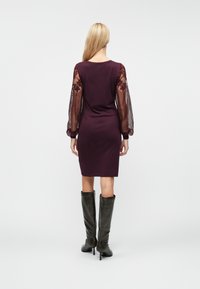 Μακρυμάνικο μωβ φόρεμα με διαφανείς δαντελένιες λεπτομέρειες στα μανίκια, εφαρμοστό σχήμα και ύψος μέχρι το γόνατο. Συνδυάζεται με ψηλές, σκούρες δερμάτινες μπότες.