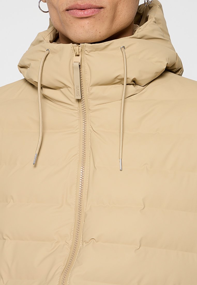 Veste beige rembourrée avec un col haut et une capuche amovible. Présente une fermeture éclair avant et des accents à cordon de serrage avec des extrémités métalliques. Texture lisse.