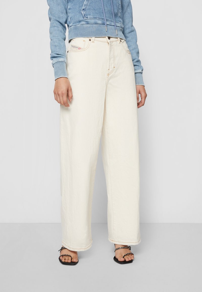 Personne portant un jean large taille haute couleur ivoire, un sweat à capuche court bleu clair et des sandales noires à fines lanières, debout sur un sol uni.