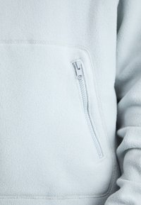Pullover in pile azzurro chiaro con tasca con zip sul lato sinistro, texture morbida e cuciture senza soluzione di continuità lungo i bordi.