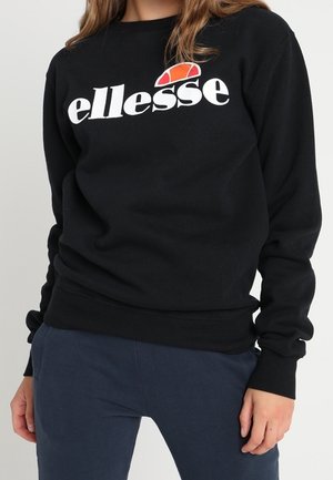 Kobieta w czarnej bluzie z białym logo "ellesse" i pomarańczową grafiką, w połączeniu z ciemnoniebieskimi spodniami, stoi z rozluźnionymi rękami.