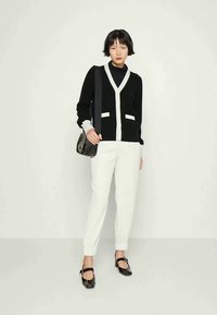 Cardigã preto e branco com decote em V, dois bolsos frontais e punhos em contraste, combinado com calças brancas ajustadas e sapatos pretos.