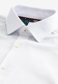Weißes Hemd mit Knopfleiste und strukturiertem Muster. Verfügt über einen klassischen Kragen und weiße Knöpfe. Das Innere hat ein florales Muster und rote Akzente.