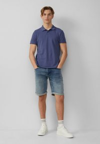 Fioletowa koszulka polo, krótkie dżinsowe spodenki z mankietami, białe sneakersy i w paski skarpetki. Model stoi z rękami w kieszeniach na jednolitym tle.