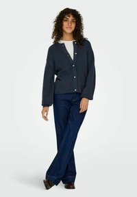 Cardigan en tricot surdimensionné bleu foncé avec un col en V, des boutons sur le devant et des manches longues, associé à un pantalon droit bleu marine et des chaussures marron.