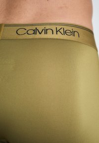 Calvin Klein vyriškos apatinės kelnės olivinės žalios spalvos, pasižyminčios lygiu paviršiumi ir juodu logotipu ant juosmens. Laikantis elegantiško, prigludusio dizaino.