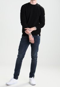 Zwarte katoenen trui met lange mouwen, gecombineerd met donkerblauwe jeans en witte sneakers. Opvallende tatoeages op de handen, casual staand.