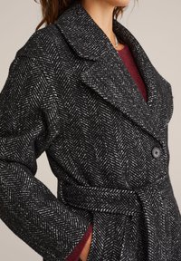 Plašč v črno-beli ribji kost z širokim ovratnikom, enojnimi gumbi in pasom za vezanje, izdelan iz teksturiranega materiala.